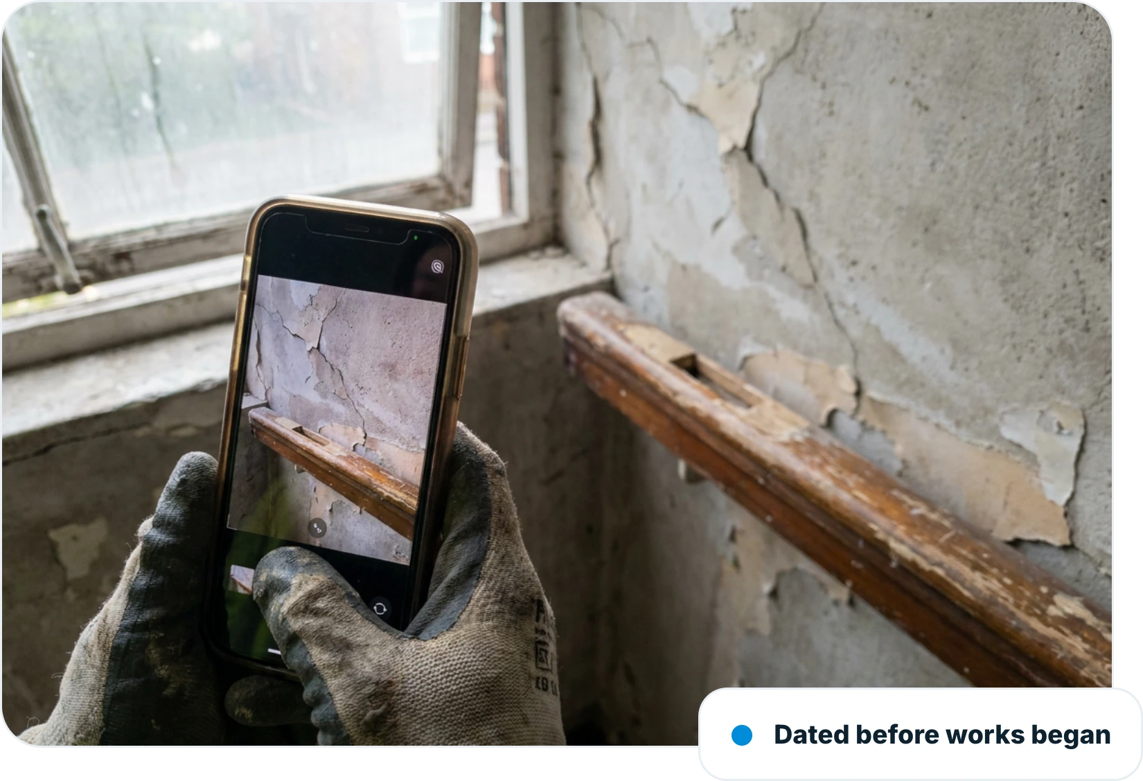 Gloved hand photographing peeling plaster and a damaged timber handrail on a phone in an empty property