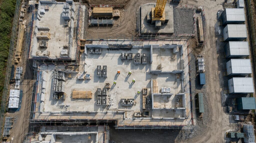 Aerial view looking straight down on a UK construction site, showing concrete slabs, stacked blockwork and timber, scaffold lines and site cabins, with workers casting long shadows.